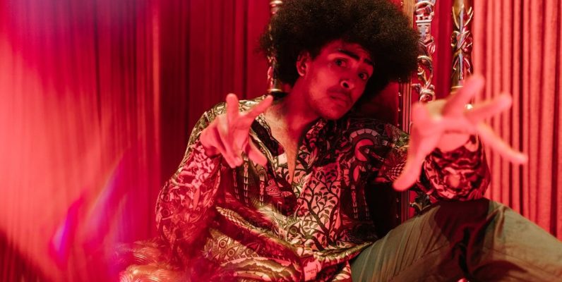 Man in vibrant outfit and afro hairstyle posing in a creative indoor setting.